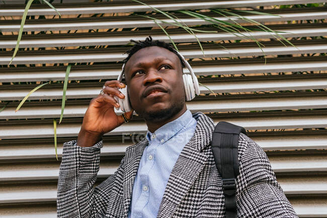 Vista lateral do jovem trabalhador de escritório afro-americano consciente ouvindo música em fone de ouvido sem fio perto da parede com nervuras na cidade — Fotografia de Stock