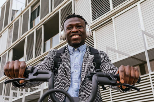 Junge zufriedene afroamerikanische männliche Angestellte im Mantel mit Fahrrad steht auf städtischem Bürgersteig gegen gerippte Wand — Stockfoto