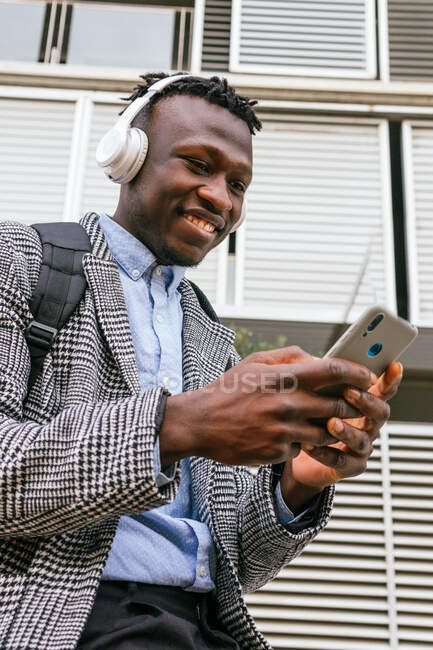 Side view of young mindful African American male office worker listening to song in wireless headset near ribbed wall in town — Stock Photo