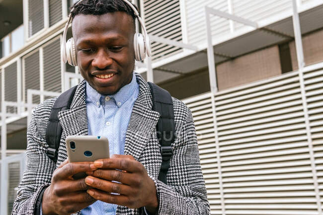Front view of young mindful African American male office worker listening to song in wireless headset near ribbed wall in town — Stock Photo