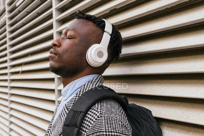 Vista lateral de un joven trabajador de oficina afroamericano consciente escuchando una canción con los ojos cerrados en auriculares inalámbricos cerca de la pared acanalada en la ciudad - foto de stock