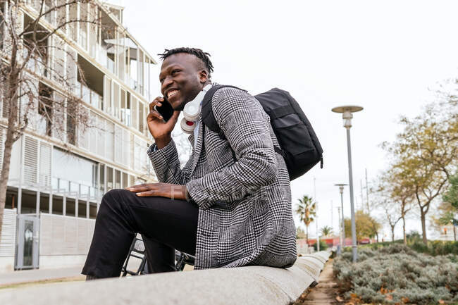 Side view of young happy African American office worker with backpack speaking on cellphone while sitting against urban building and looking away — Stock Photo