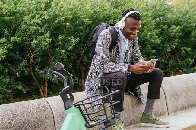 Молодий позитивний етнічний чоловічий офіс співробітник в навушниках спілкується на мобільному телефоні, відпочиваючи біля велосипеда в місті — стокове фото