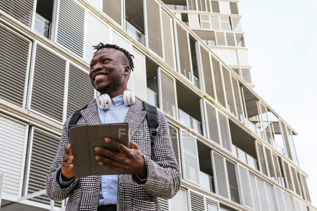 From below of young glad African American male manager with tablet and headset looking away against urban building — Stock Photo