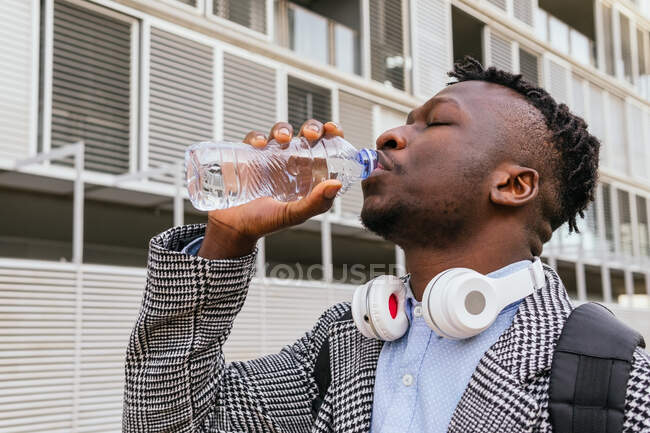 Young African American male manager with headphones and closed eyes drinking water from bottle against building in town — Stock Photo