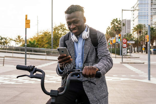 Jovem trabalhador de escritório preto feliz com fone de ouvido conversando no celular enquanto sentado em bicicleta no pavimento urbano — Fotografia de Stock