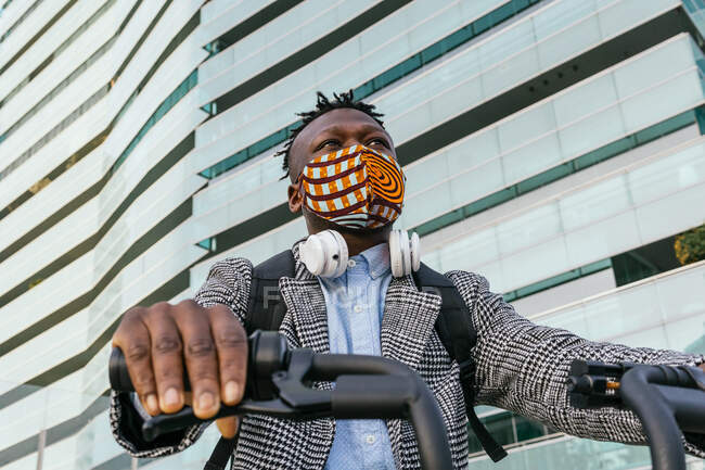 Low angle of anonymous ethnic male manager in mask and formal wear riding bicycle against urban buildings while looking away — Stock Photo