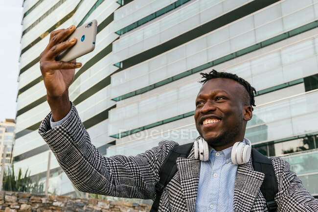 Junge positive männliche Büroangestellte mit Kopfhörern, die bei Tageslicht ein Selbstporträt auf dem Handy gegen städtische Gebäude machen — Stockfoto