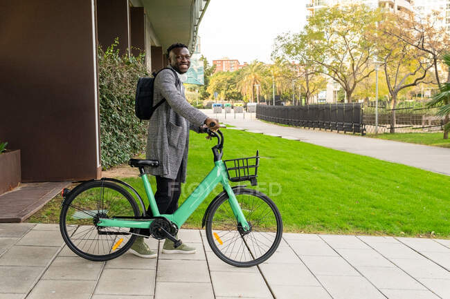 Вид сбоку на молодого счастливого этнического менеджера с рюкзаком и велосипедом, смотрящего в камеру на городское здание — стоковое фото