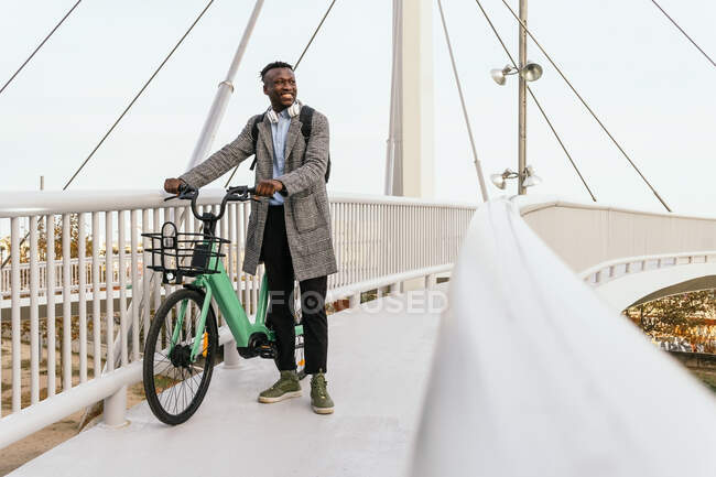 Young smiling ethnic male office worker in formal outfit with headphones and bicycle looking away on suspension bridge in city — Stock Photo