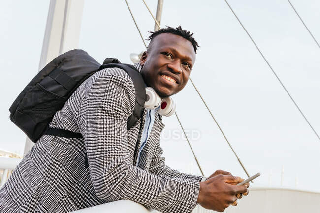 Vista lateral de un joven y alegre gerente afroamericano con mochila y teléfono celular apoyado en la cerca del puente urbano mientras mira hacia otro lado - foto de stock