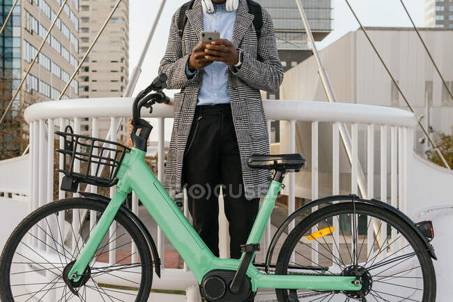 Baixo ângulo de conteúdo anônimo Gerenciador masculino afro-americano em mensagens de texto de fone de ouvido no celular perto de bicicleta na cidade — Fotografia de Stock