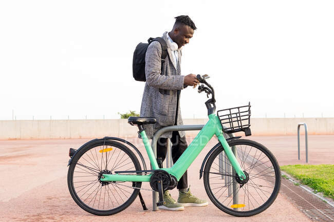 Бічний вид афроамериканського працівника чоловічої статі, який користується додатком на мобільному телефоні під час паркування велосипеда в місті. — стокове фото