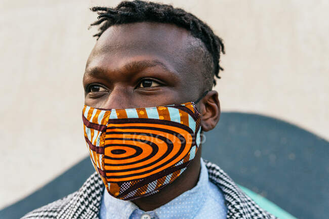 Anonyme kontemplative ethnische männliche Manager in dekorativer Maske mit Handy in der Nähe der Stadtmauer während der COVID-19-Pandemie — Stockfoto