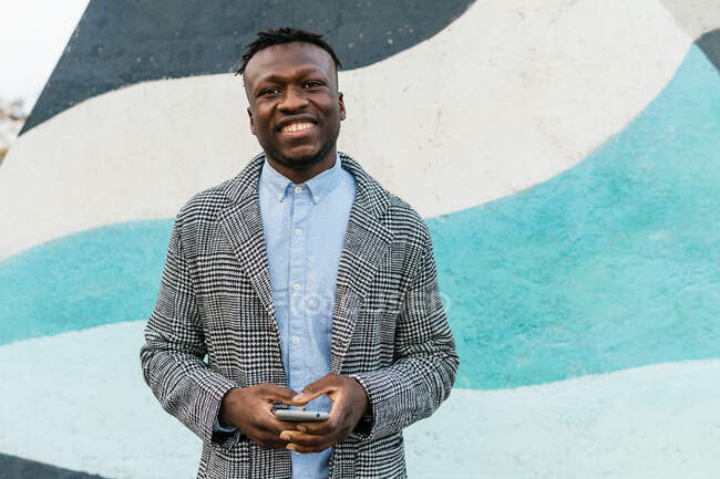 Junger positiver schwarzer männlicher Manager in formeller Kleidung mit modernem drahtlosen Headset, der in der Nähe einer hellen Mauer in die Kamera blickt — Stockfoto