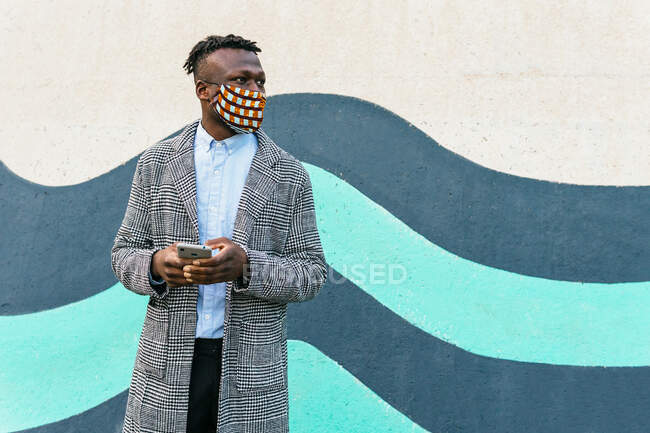 Anonymous contemplative ethnic male manager in ornamental mask with cellphone looking away near urban wall during COVID 19 pandemic — Stock Photo