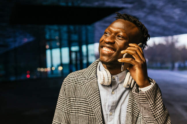 Jovem sorrindo étnico masculino trabalhador de escritório em fones de ouvido falando no celular enquanto olha para longe na rua à noite — Fotografia de Stock
