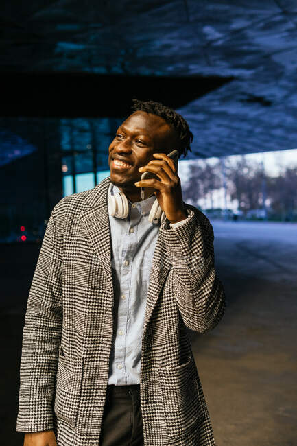 Молодой улыбающийся работник офиса этнического мужчины в наушниках, разговаривающий по мобильному телефону, глядя на улицу вечером — стоковое фото