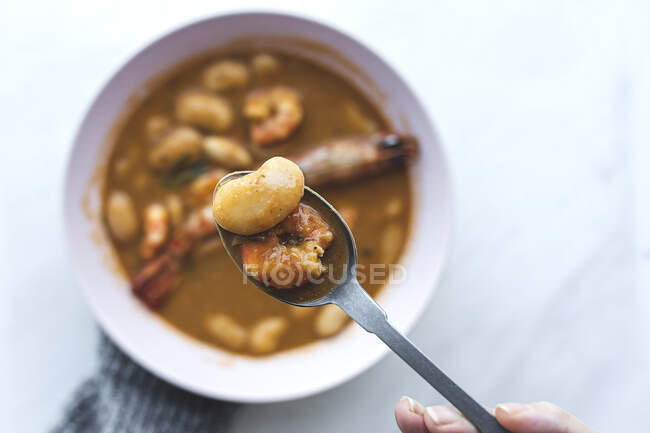 Top view of crop anonymous person in warm sweater sitting at white marble table and eating delicious soup with seafood and white beans served in bowl — Stock Photo