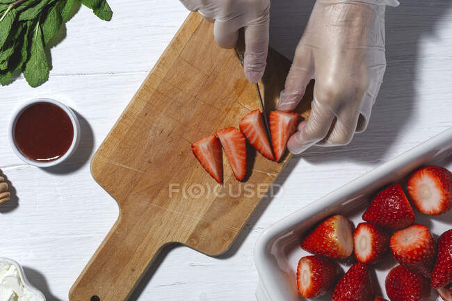 Top vista cultura chef irreconhecível em luvas de látex cortar morangos deliciosos frescos na tábua de corte de madeira na mesa — Fotografia de Stock