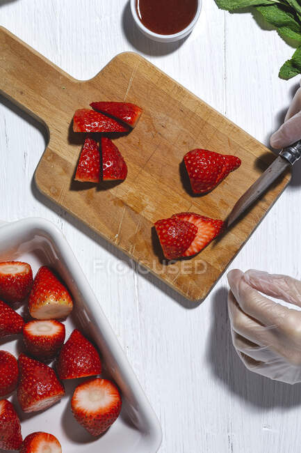 Top view cosecha irreconocible chef en guantes de látex corte fresas frescas deliciosas en tabla de cortar de madera en la mesa - foto de stock