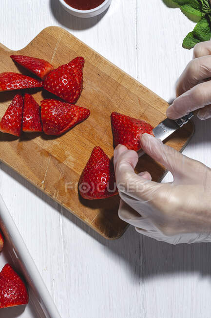 Top view raccolto chef irriconoscibile in guanti di lattice taglio fresco deliziose fragole sul tagliere di legno sul tavolo — Foto stock