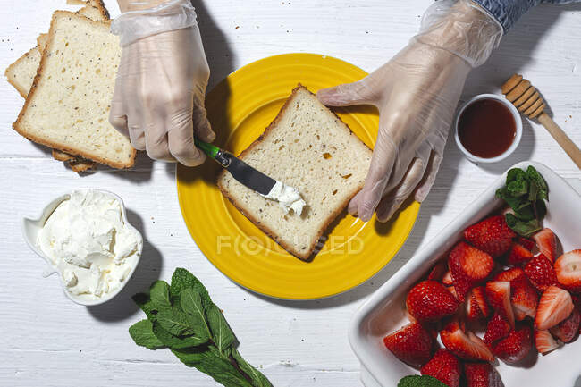 Top view raccolto anonimo femminile cuoco in guanti di lattice diffusione gustosa crema di formaggio sulla fetta di pane posto sul piatto giallo vicino a fragole tagliate — Foto stock