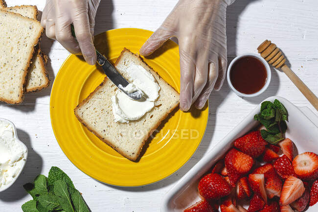 Vue de dessus récolte cuisinière anonyme dans des gants de latex étalant fromage à la crème délicieux sur tranche de pain placé sur plaque jaune près de fraises coupées — Photo de stock