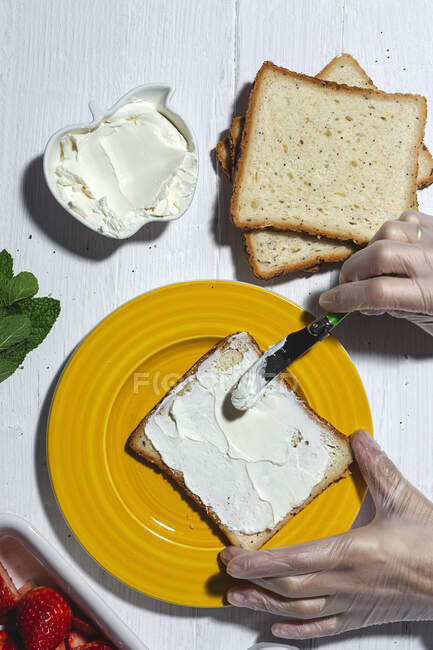 Анонимная кухарка в латексных перчатках, разбрасывающая вкусный сливочный сыр на ломтик хлеба, помещенный на желтую тарелку — стоковое фото