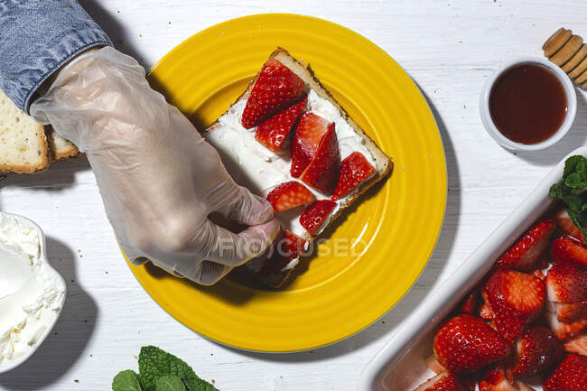 Top view crop anonymous chef in glove arranging cut strawberries on bread toast with spread cream cheese — Stock Photo