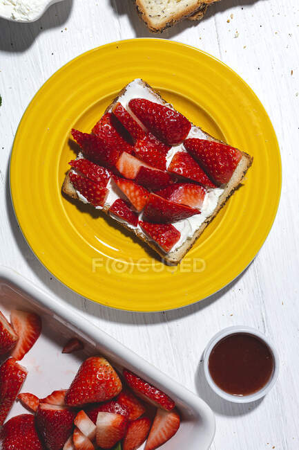Накладний склад солодкого тосту з вершковим сиром і стиглою полуницею, подається на жовтій тарілці на столі — стокове фото