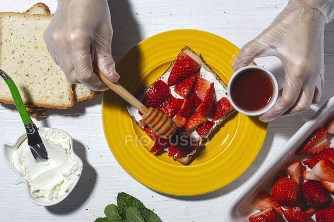 Overhead crop anonymous skilled female chef in gloves pouring sweet honey on delicious toast with cream cheese and cut strawberries while cooking in light kitchen — Stock Photo