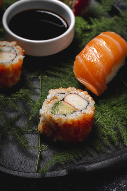 Gustoso sushi fresco assortito servito su ramoscelli di piante verdi su piatto nero con salsa di soia sul tavolo di marmo — Foto stock