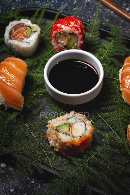 Tasty fresh assorted sushi served on green plant twigs on black plate with soy sauce on marble table near chopsticks — Stock Photo