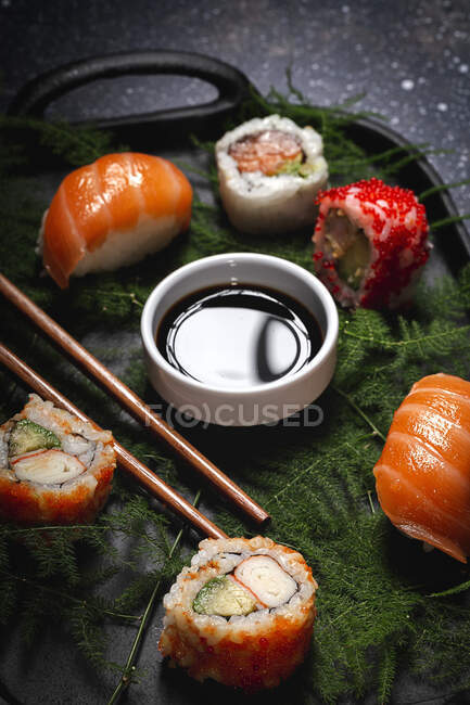 Sabroso sushi fresco servido en ramitas de plantas verdes en plato negro con salsa de soja en mesa de mármol cerca de palillos - foto de stock