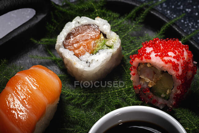 Tasty fresh assorted sushi served on green plant twigs on black plate with soy sauce on marble table near chopsticks — Stock Photo