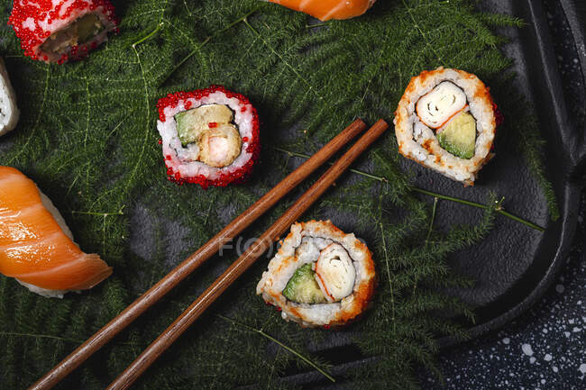 Вкусные свежие суши подаются на зеленых ветках растений на черной тарелке с соевым соусом на мраморном столе рядом с палочками для еды — стоковое фото
