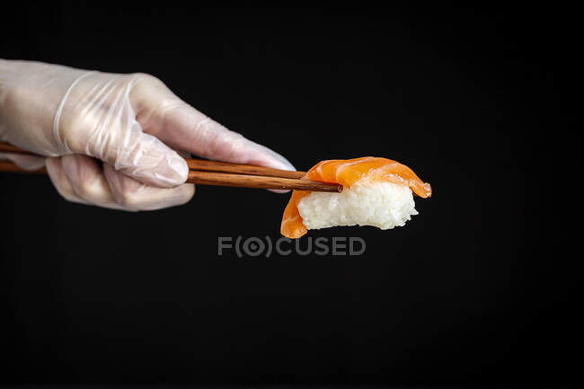 Crop anonymous chef in latex glove holding salmon sushi with chopsticks against black background in modern Japanese restaurant — Stock Photo