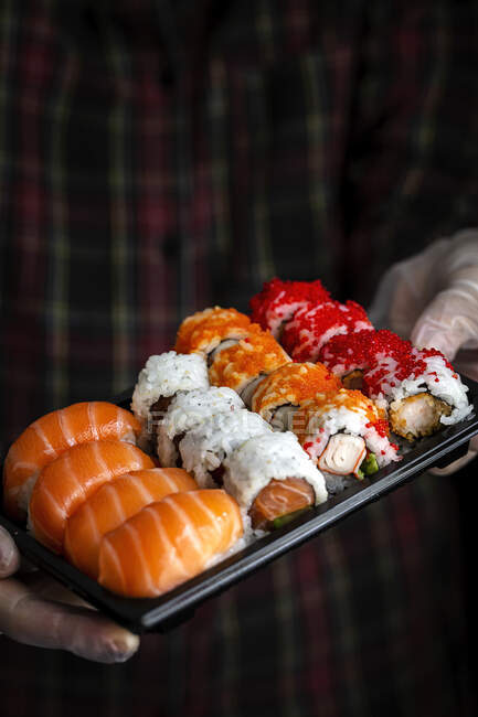 Crop unrecognizable chef in gloves showing platter with set of palatable assorted sushi in dark room — Stock Photo