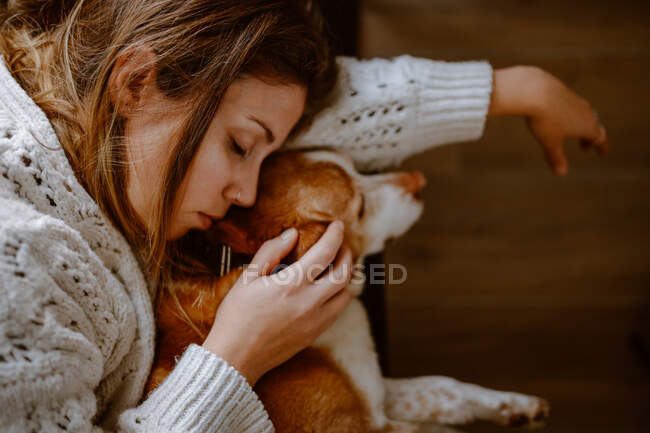 Vista superior de la cosecha joven hembra en suéter cálido abrazando adorable perro de raza pura mientras duermen juntos en el sofá en casa - foto de stock