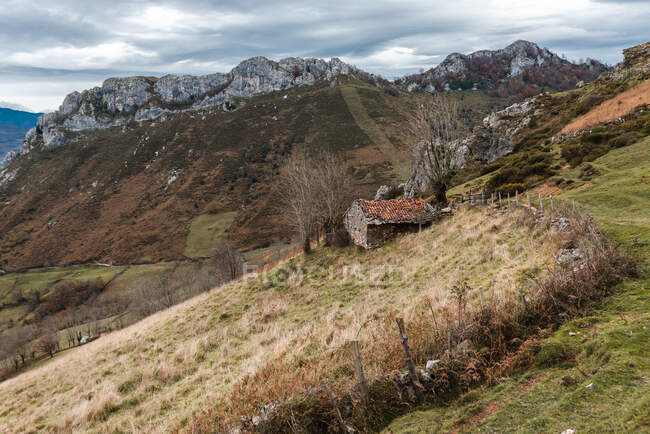 Casa rurale abbandonata intemperie vicino al sentiero sul pendio erboso in un ampio terreno montagnoso nella giornata nuvolosa nelle Asturie Spagna — Foto stock