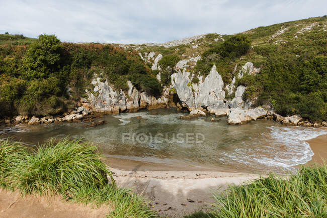Picturesque view of small sandy shore of rippling foamy seawater surrounded by rough cliffs covered with lush greenery on clear summer day in Asturias — Stock Photo