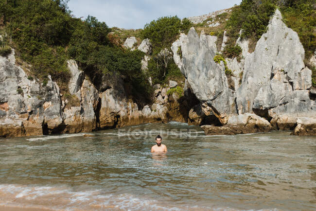 Hombre delgado sin vestir parado en la tranquila playa interior del lago rodeado de escarpados acantilados rocosos en Asturias - foto de stock