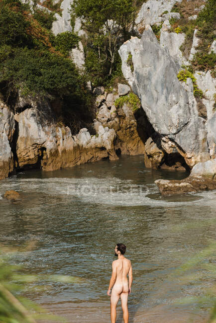 Back view anonymous undressed slim male standing on tranquil lake inland beach surrounded by rough rocky cliffs in Asturias — Stock Photo