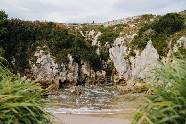 Malerischer Blick auf die Ferne unkenntlich Person, die oben auf dem Berghang in der Nähe von kleinen sandigen Ufer von schäumendem Meerwasser mit üppigem Grün an klaren Sommertagen in Asturien bedeckt — Stockfoto