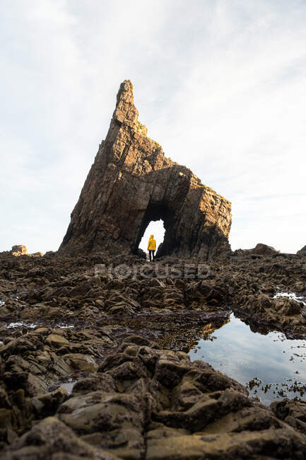 Далекий путешественник в теплой куртке, стоящий на естественной арке грубой скалы, расположенной на каменистом огромном побережье в ясный осенний день — стоковое фото