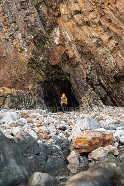 Back view anonymous traveler in warm yellow jacket standing near rough rock grotto entrance located on rocky stony terrain — Stock Photo