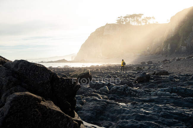 Viajante anônimo em casaco amarelo quente andando em litoral de pedra áspera cercado por penhascos severos na luz solar — Fotografia de Stock