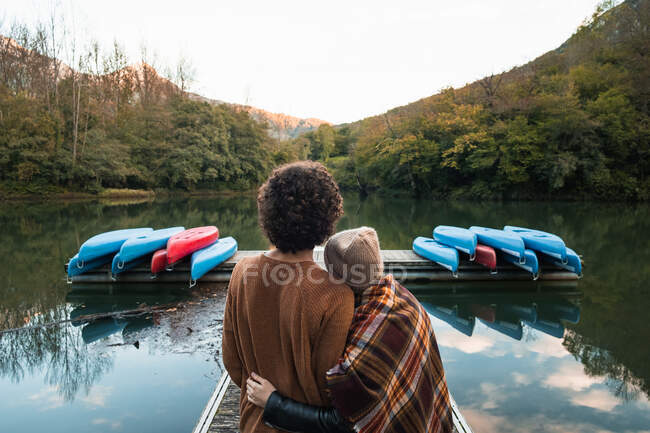 Back view young loving couple in warm wear embracing each other tenderly while standing on lake wooden quay against lush abundant forested hills in reservoir Valdemurio — Stock Photo