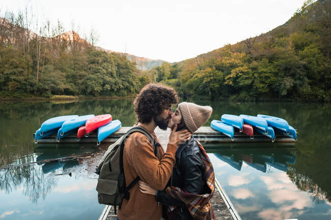Side view young loving couple in warm wear kissing tenderly while standing on lake wooden quay against lush abundant forested hills in reservoir Valdemurio — Stock Photo